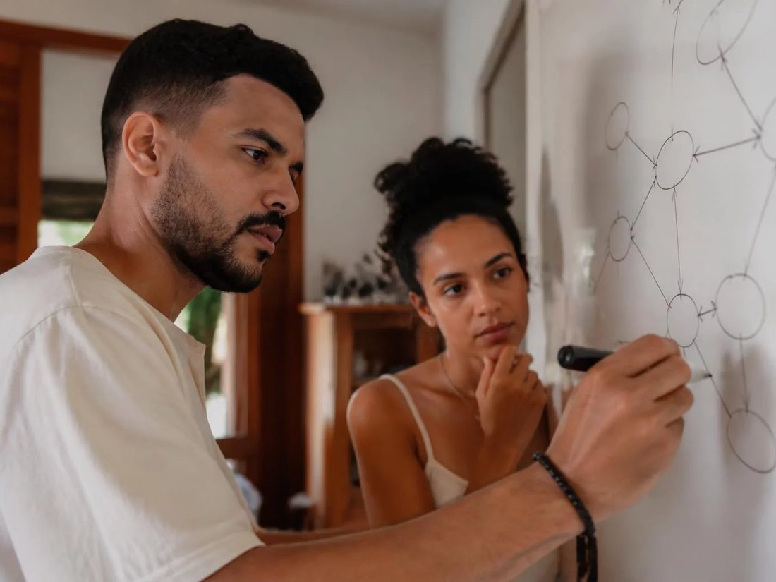 Dupla em pé diante de um quadro branco com o esqueleto de um argumento desenhado
